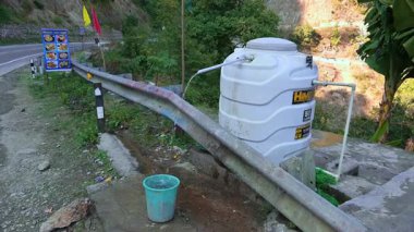 Rishikesh, Hindistan - 08 Ocak 2025: Himalaya yolunun yakınındaki bir kovayı bir musluk dolduruyor. Yakınlarda bir restoran panosu kahvaltı, öğle yemeği ve akşam yemeği için menü sunar..