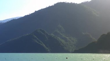 Durgun bir manzara, mavi göldeki su dalgalarını sergiliyor. Çerçevelenmiş dağlar, yemyeşil ağaçlarla bezenmiş..