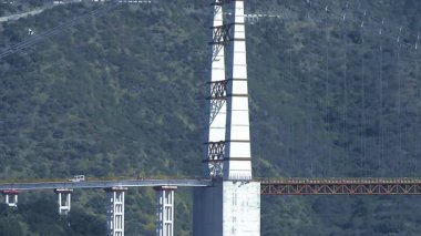 Canlı bir sahne, insanlar yakınlarda dururken ve dağlar dramatik bir zemin oluştururken köprüden geçen trafiği resmediyor..