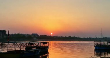 Mathura 'daki Yamuna Nehri' ndeki evlerin arkasındaki günbatımı, teknelerin kenetlendiği ve suyun rengarenk yansımasıyla çarpıcı bir akşam manzarası yaratıyor..