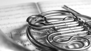 Siyah ve beyaz, nota kağıtlarının üzerinde dinlenen Fransız kornosunun yakın plan çekimi. Yavaş kamera hareketi. Proffesional orkestra veya bando oyuncuları tarafından kullanılan parlak borulardan yapılmış bir pirinç enstrüman..