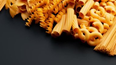 Makarna yığınının kusursuz bir animasyonu. Koyu yüzeyde farklı türde çiğ makarna var. Makarna, rigatoni, farfalle, spagetti, makarna. İtalyan mutfağı. 4k HD