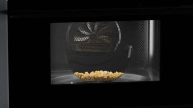 Mikro dalgada dönen cam kaseden çıkan mısır tohumlarının animasyonu. Evde film izlerken lezzetli tuzlu abur cuburlar yemek için hazır. Patlamış mısır hazırlığı. Siyah bir fırın. Karanlık arka plan. 4K