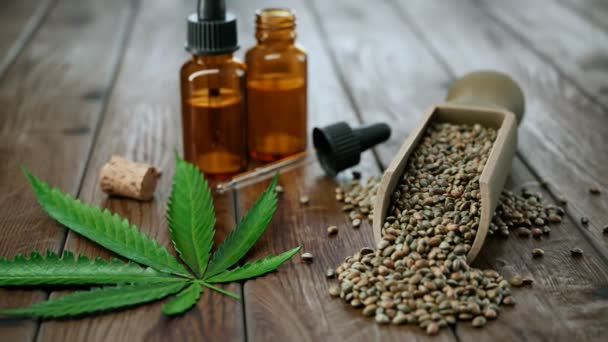 テーブルの上の大麻オイルとのアニメーション 木のスプーンでマリファナの緑の葉と麻の種子の隣の小さなガラス瓶のCbdエッセンシャルオイル 大麻の治療および化粧品の使用の概念 — ストック動画