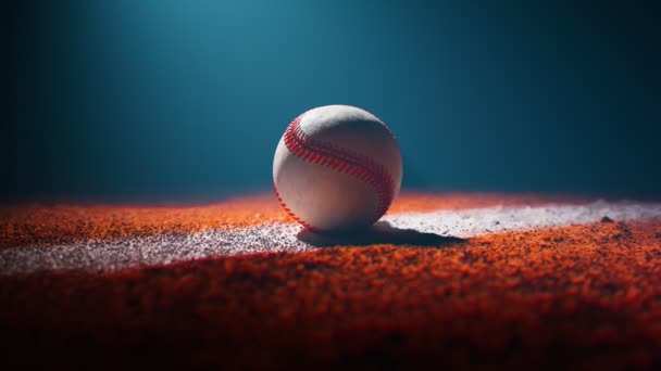 棒球躺在涂满橙色泥土的白色田边上 黑暗的体育场 只有一个聚光灯照明球 美国国家体育装备在地面上 相机绕着它旋转 — 图库视频影像