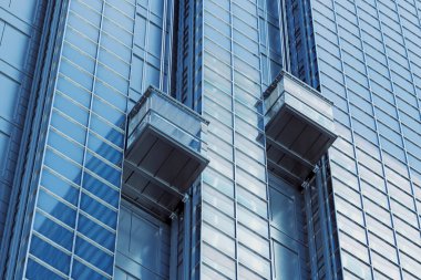 Modern bir gökdelende iki asansör yukarı çıkıyor. Mavi gökyüzü pencerelere yansıyor. Bina, modern mimari ve tasarımın sergilendiği hareketli bir şehir.