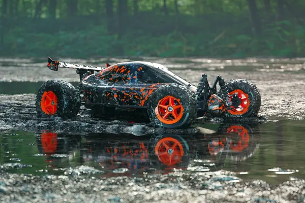 Dirty, orange RC-Car got stuck in the mud on a wet forest mire path. Green, lush trees and car covered with mud reflected in a big puddle of rainwater. Lost race. Last place. Broken toy.