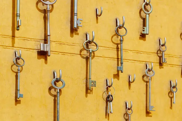 Various sizes and types of keys hanging on hooks on the yellow wall. The keys create a unique visual display that grab the attention of any viewer. Showing signs of wear and tear from years of use