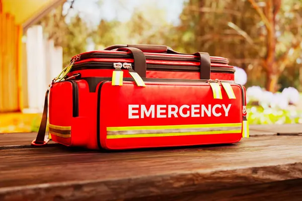Professional red emergency first aid bag on a wooden table outdoors. Doctor\'s kit stacked with equipment and ready for fast response in a green, lush, natural environment. Saving lives.