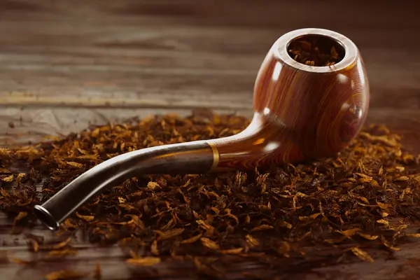 A classy wooden pipe with intricate golden details rests on a pile of scattered tobacco on a vintage wooden table. Elegance and style of the tobacco culture, with its rich history and timeless appeal.