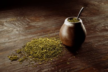Geleneksel Yerba Mate içki setinin yakın plan çekiminde ahşap bir su kabağı, bombilla kamışı ve doğal ahşap arka planda dağınık eş yaprakları yer alır..
