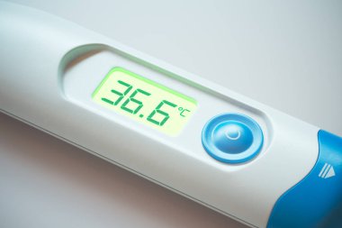 Dijital termometrede yüksek çözünürlüklü yakın çekim ve 36.5C değerleri klinik arka planda normal vücut ısısını gösteriyor.