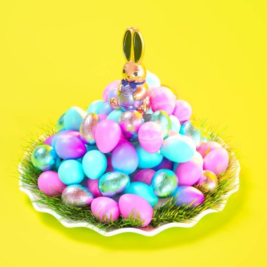 Bu nefis Paskalya düzeninde, yumuşak çimenlere yerleştirilmiş renkli yumurta çeşitlerini sunan, sevimli bir tavşancık süsü ile tamamlanmış, canlı sarı renkli bir renge karşı.