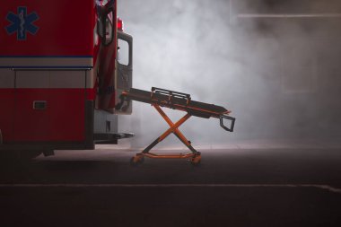 Bu resimde, sis yüklü bir atmosfer acil durum sahnesini gizliyor, acil tıbbi müdahaleye hazır bir ambulans ve sedye ile..