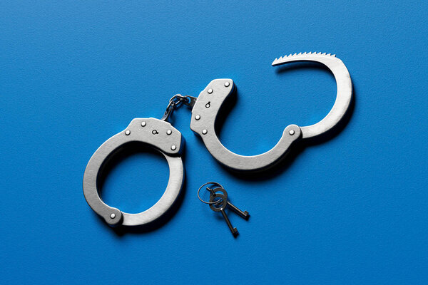 A detailed view of a pair of open metal handcuffs with accompanying keys sprawled on a striking blue background, encapsulating themes of security, legal authority, and control.