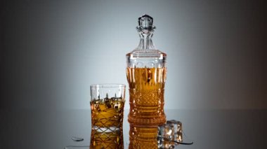 Lüks bir kristal sürahi ve yarısı kehribar renkli bir ruhla dolu, gri bir zemin üzerine kurulmuş, yansıtıcı yüzeyinde buz küpleri olan bir cam..