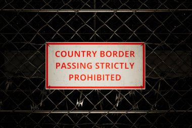 Intense close-up view of a stark warning sign atop a secure chain-link fence delineating a national border, symbolizing strict regulations and enforced prohibition of unauthorized border crossing. clipart