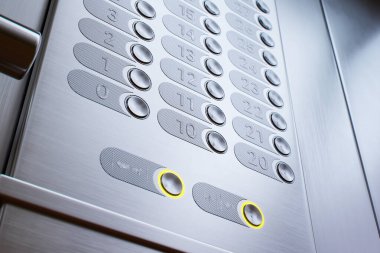 Şık bir asansör kontrol panelinin detaylı bir görüntüsü, sıfır ve birinci katların aydınlatma düğmelerini gösteriyor, gelişmiş erişilebilirlik için dokunulabilirlik numaralarıyla fırçalanmış metal bir kaplama içinde..