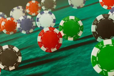Kumarhane poker çiplerinin dinamik ve renkli dizisi yeşil ahşap bir yüzeye saçılmış vaziyette duruyor. Bu da kumar ortamı ve oyun stratejisini canlandırıyor..