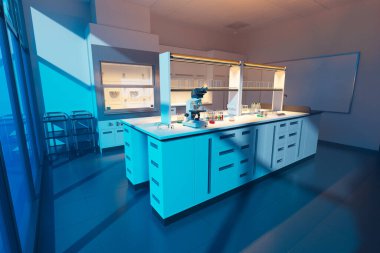 Bu resim modern bir laboratuvar iç mekanını çekiyor, mikroskoplar, bilimsel cihazlar ve mavi dolaplar dahil olmak üzere en son teknolojiye sahip ekipmanların hepsi akşam ışığı altında..