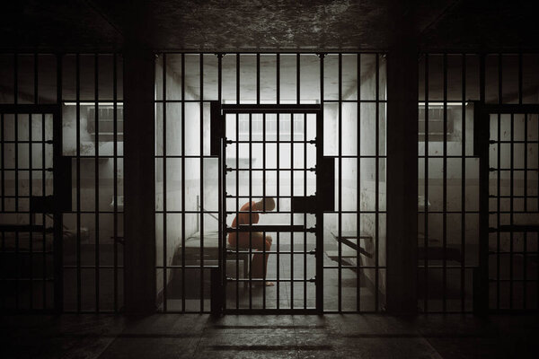 This impactful image shows the stark silhouette of a person seated alone in a dimly lit jail cell, with rays of light casting shadows through the bars.