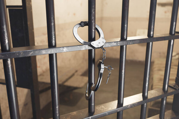 Металлические наручники свисают с темных железных прутьев тюремной камеры, воплощая понятия справедливости, правоприменения, наказания и сложности свободы.