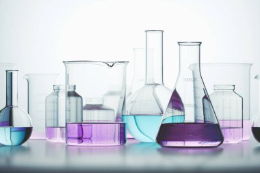 Canlı kimyasal çözümlerle dolu deney şişeleri ve şişeler de dahil olmak üzere çeşitli laboratuvar vitrinleri, bilimsel araştırma ve kimyasal analizlerin göstergesi olarak beyaz bir bankta sergilenmektedir..