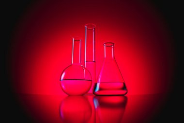 Bu resim, bilimsel araştırmaları ve deneyleri sembolize eden kırmızı bir zemine karşı canlı bir laboratuvar cam kapları koleksiyonunu sergiliyor..