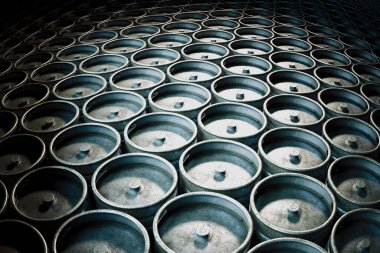 Bu görüntü endüstriyel metal fıçılardan oluşan geniş bir koleksiyon yakalıyor, loş bir depo alanında düzenli bir şekilde dizilmiş, tekdüzeliğin ve endüstriyel dokuların güzelliğini vurguluyor..