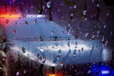 Pencerenin camındaki su damlacıklarını kapatarak gecenin renkli sokak lambaları büyüleyici ve soyut bir yansıma yaratıyor..