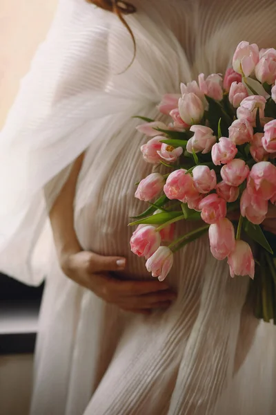 Pregnant woman in a stylish white dress holding her belly with one hand a bouquet of pink tulips in the other hand. Close up photo of pregnant belly. Stylish Motherhood concept