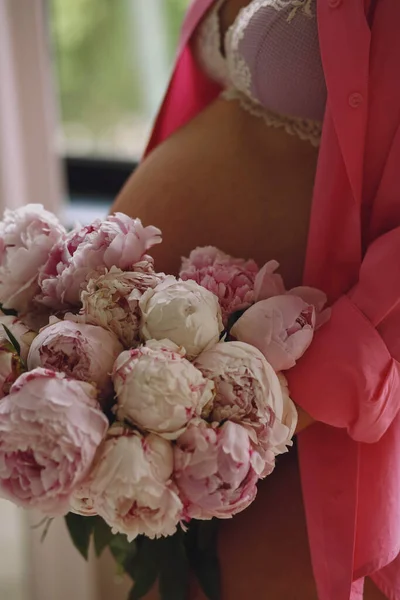 Pregnant woman in a stylish pink underwear and pink shirt holding the bouquet of pink peonies in her hands near pregnant belly. Close up photo of pregnant belly. Stylish Motherhood concept