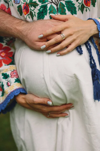 快乐的年轻孕妇穿着乌克兰传统民族风格的衣服 丈夫抱着怀孕的肚子 有着自然的背景 孕妇腹部近照 — 图库照片