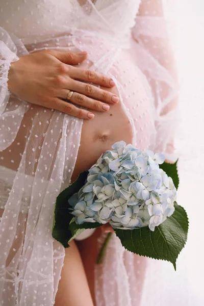 穿着时髦的白色衣服的怀孕妇女一只手牵着她的腹部 另一只手牵着蓝色的水仙花 孕妇腹部的近照 典型的母性概念 — 图库照片