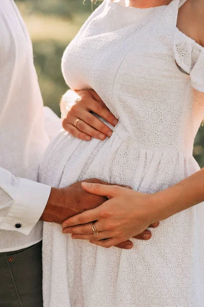身穿白色夏装的孕妇和她的丈夫在大自然的背景下手拉着怀孕的腹部 爱的概念 家庭观念快乐生儿育女的概念 — 图库照片
