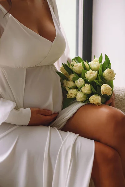 怀孕的女人穿着漂亮的白色衣服 一只手拿着一束黄色的郁金香 另一只手牵着她的肚子 孕妇腹部的近照 典型的母性概念 — 图库照片