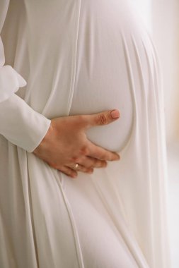 Hamile kadın, karnını tutan beyaz bir elbise. Hamile karnının yakın fotoğrafını çek. Annelik kavramı