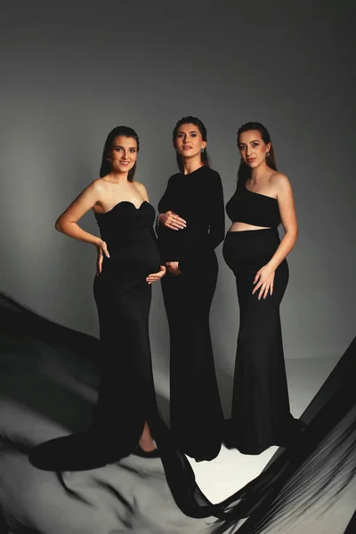 Happy pregnant women in black dresses with big bellies. Group of positive pregnant women wearing black dresses and posing in studio with black fabrick. Happy Motherhhood