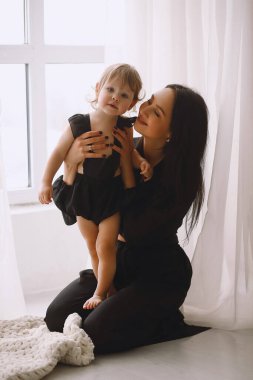 Siyah elbiseli anne pencerenin yanında oturup iki yaşındaki kızına sarılıyor. Mutlu anne, küçük kızına evde sarılıyor. Anne ve kızı.