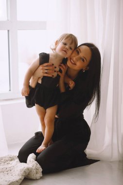 Siyah elbiseli anne pencerenin yanında oturuyor ve iki yaşındaki kızına yumuşak bir şekilde sarılıyor. Mutlu anne, küçük kızına evde sarılıyor. Anne ve kızı.