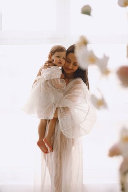 Büyük pencerenin yanında durup bebeğine sarılan beyaz elbiseli mutlu annenin stüdyo portresi. Beyaz elbiseli bir anne ve kız. Mutlu annelik kavramı.