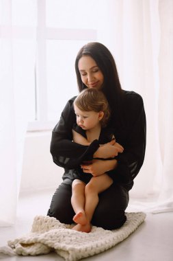 Siyah elbiseli esmer kadın pencerenin yanında oturup iki yaşındaki kızına sarılıyor. Mutlu anne, küçük kızına evde sarılıyor. Anne ve kızı.