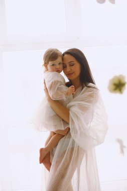 Büyük pencerenin yanında duran ve iki yaşındaki kızına sarılan beyaz elbiseli mutlu annenin stüdyo portresi. Beyaz elbiseli bir anne ve kız. Mutlu annelik kavramı.