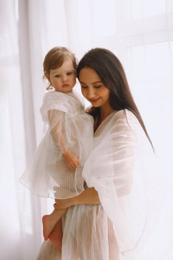 Beyaz elbiseli mutlu bir annenin stüdyo portresi büyük pencerenin yanında duruyor ve küçük kızını kollarına alıyor. Beyaz elbiseli bir anne ve kız. Mutlu annelik kavramı.