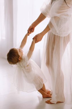 Annesi ve iki yaşındaki beyaz elbiseli kızı stüdyoda büyük pencerenin yanında oynuyorlar. Kızıma bakıyorum. Kızların Günü. Metin için alanı kopyala