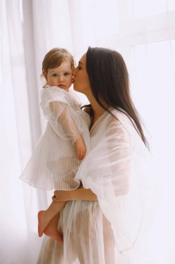 Beyaz elbiseli mutlu bir annenin stüdyo portresi büyük pencerenin yanında duruyor ve küçük kızını kollarına alıp öpüyor. Beyaz elbiseli bir anne ve kız. Mutlu annelik kavramı.