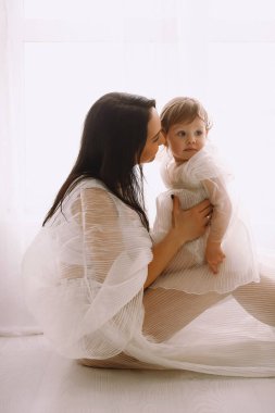 Beyaz elbiseli mutlu bir annenin stüdyo portresi. Pencerenin yanında oturmuş bebeğini kucağında tutuyor. Beyaz elbiseli anne ve kız. Mutlu annelik ve çocukluk kavramı.
