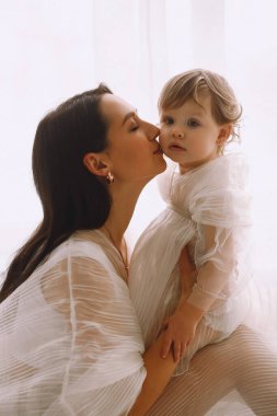 Beyaz elbiseli mutlu bir annenin, pencerenin kenarında oturup bebeğini kollarına alıp öptüğü yakın bir fotoğraf. Beyaz elbiseli anne ve kız. Mutlu annelik ve çocukluk kavramı.