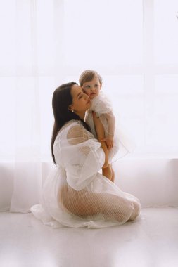 Beyaz elbiseli annenin stüdyo resmi. Pencerenin yanında oturmuş bebeğine sarılıyor. Beyaz elbiseli anne ve kız. Mutlu annelik ve çocukluk kavramı.