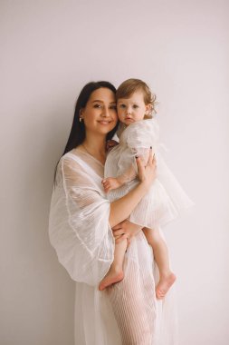 Beyaz elbiseli mutlu bir annenin stüdyo portresi duvarın yanında duruyor ve küçük kızını kollarına alıyor. Beyaz elbiseli bir anne ve kız. Mutlu annelik kavramı.